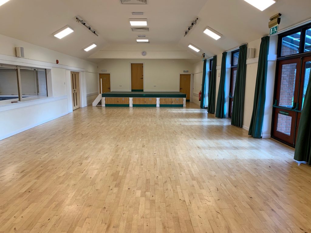 Spacious interior of Assington Village Hall featuring a bright, open layout with polished wooden floors, large windows letting in natural light