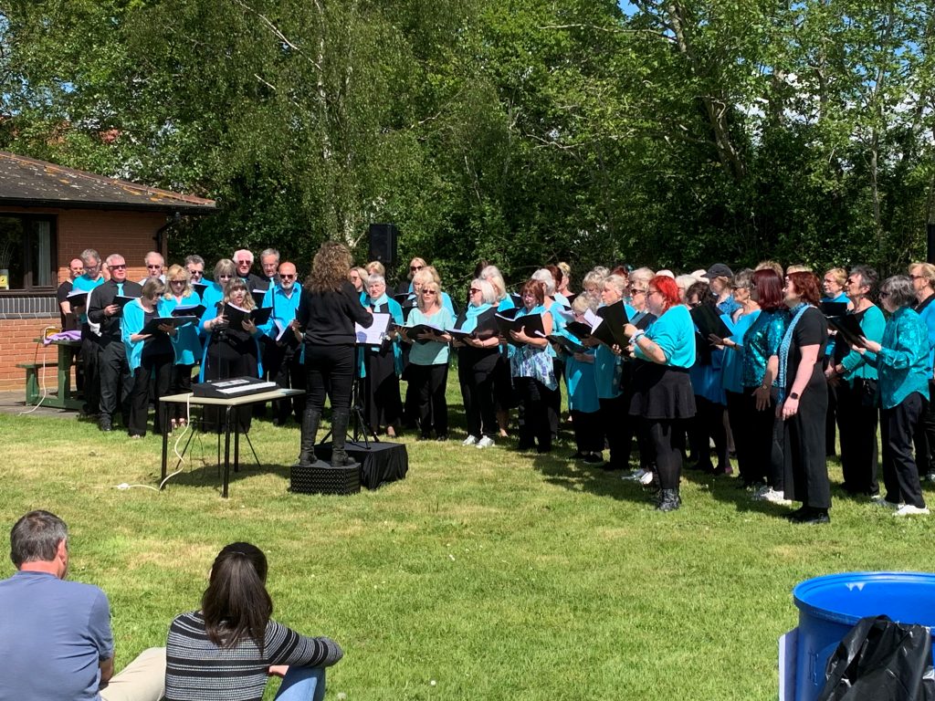 Happy Sounds Choir performing outdoors on a sunny day, led by a conductor, with an audience watching. The community choir regularly hires Assington Hall for rehearsals and events.